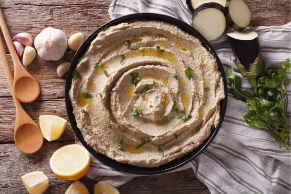 bowl of eggplant baba ganoush with garlic cloves, lemon and sliced eggplant